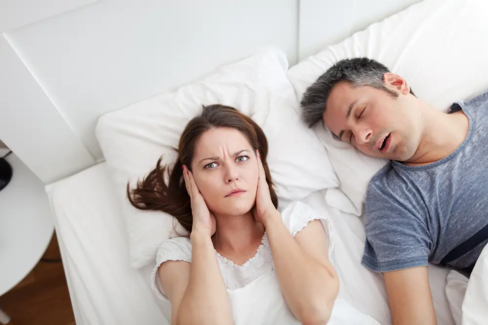 A man snoring next to a woman covering her ears