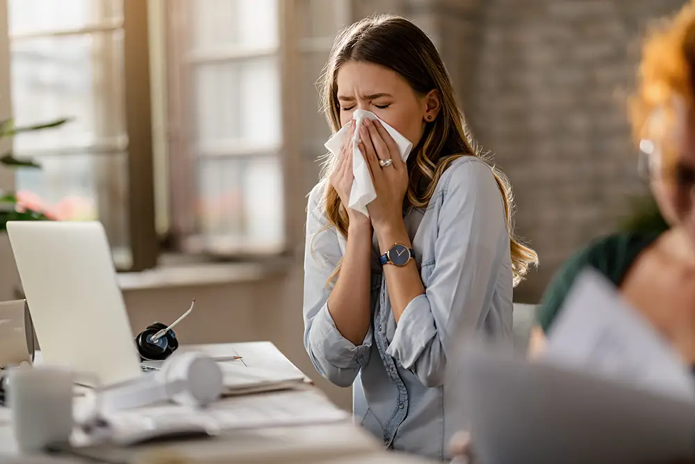 A photo of a woman sneezing into a tissue at her work desk, illustrating the need for allergy treatment